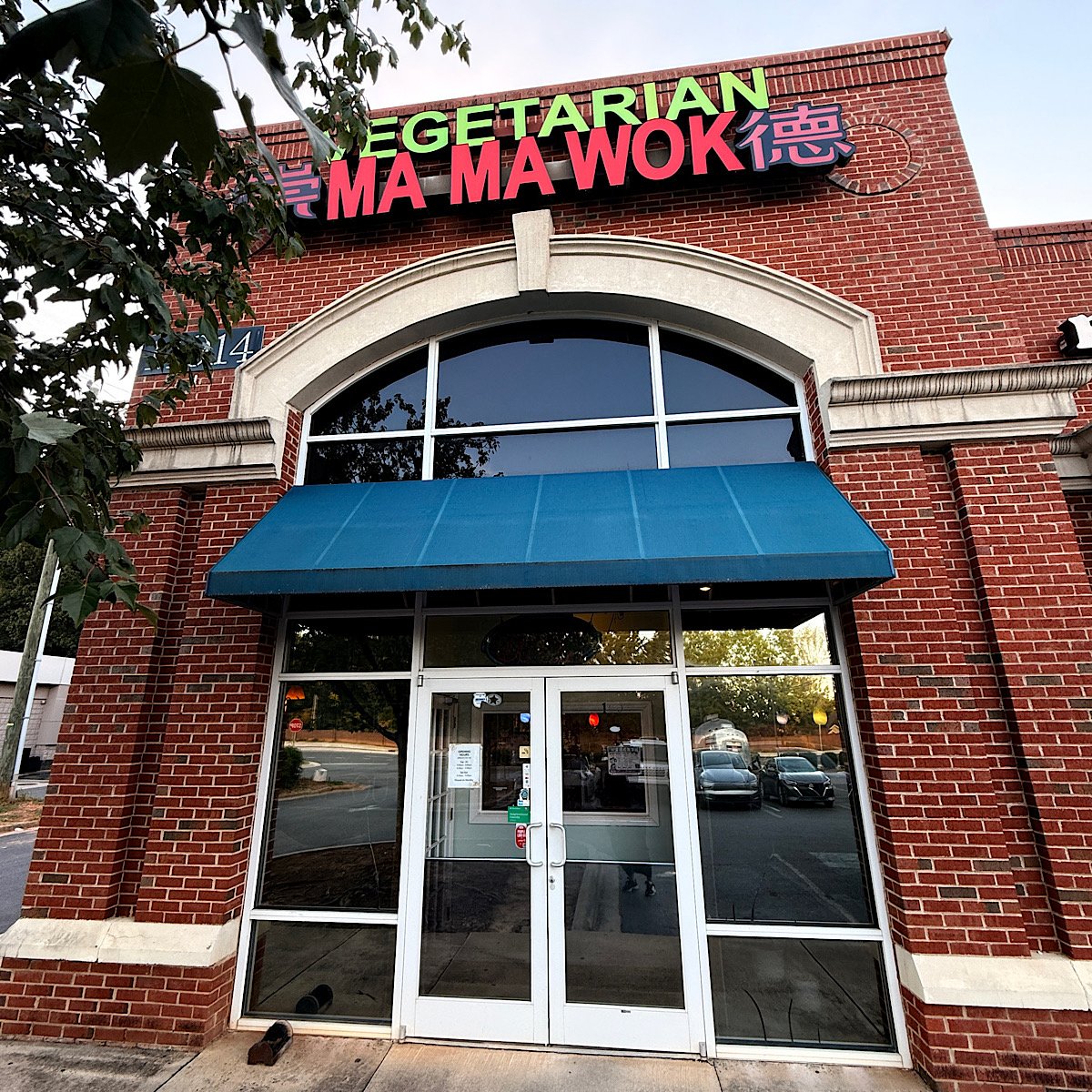The entrance of a brick restaurant building with a blue awning and glass doors, featuring a neon sign that reads Vegetarian Ma Ma Wok in English and Chinese characters.