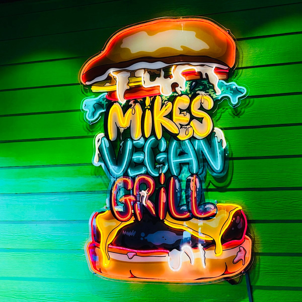 A colorful neon sign shaped like a burger on a green wood wall reads Mikes Vegan Grill with dripping letters and vibrant accents, suggesting a lively, plant-based restaurant.