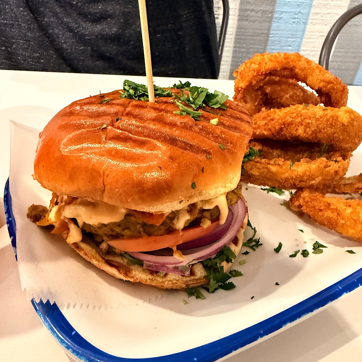 A grilled bean burger with lettuce, tomato, red onion, and sauce in a toasted bun, garnished with chopped herbs, served with crispy onion rings on a tray lined with paper.