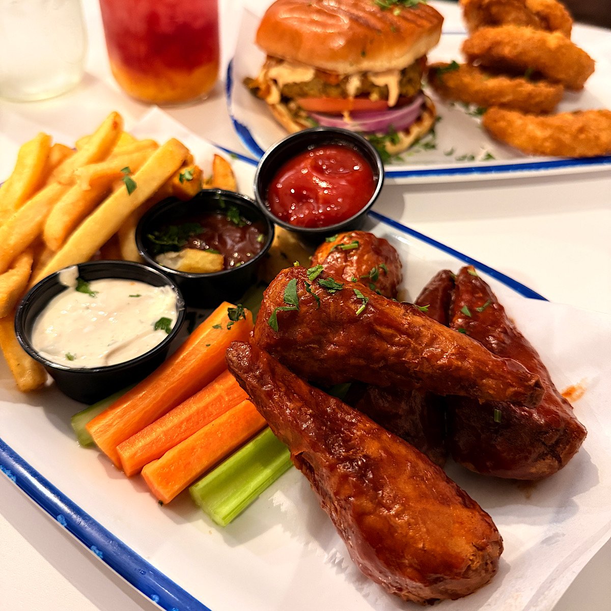 A plate with vegan sauced chicken wings, carrot and celery sticks, dipping sauces, and fries. In the background, a vegan bean burger, onion rings, and a colorful drink are visible on a white table.