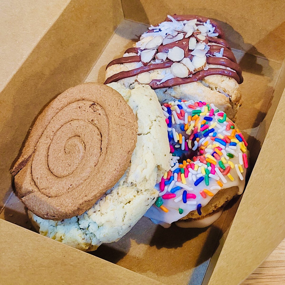 Three large vegan cookies in a brown box: one with a cinnamon swirl design, one topped with white icing and rainbow sprinkles, and one with chocolate drizzle, coconut, and almond slices.