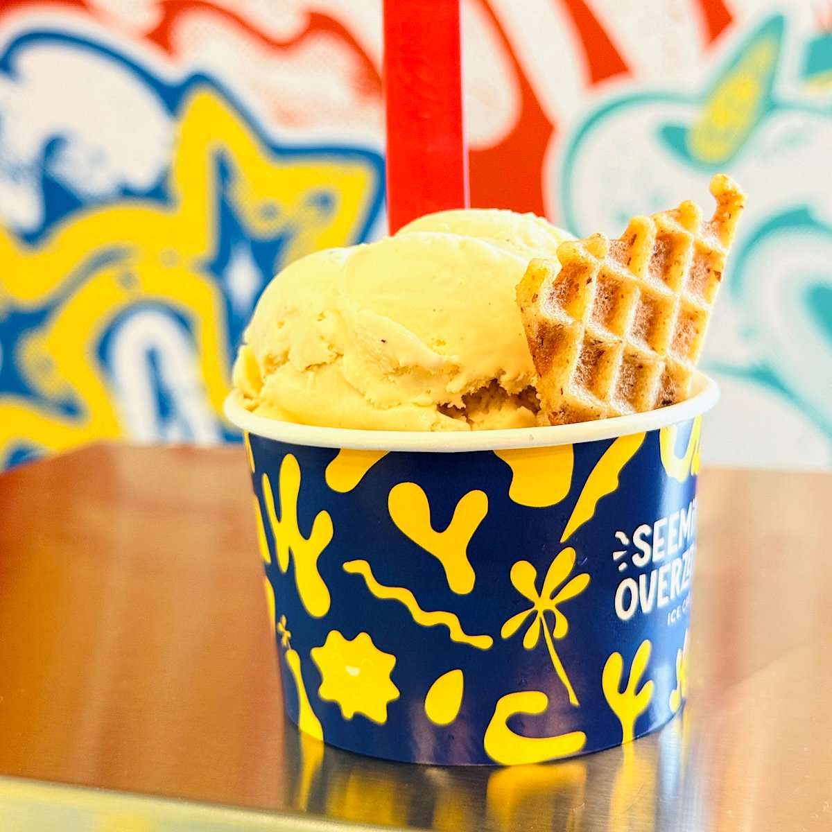 A cup of vegan ice cream with two waffle pieces, served in a blue and yellow patterned cup, sits on a reflective surface. A bright, colorful mural is blurred in the background.