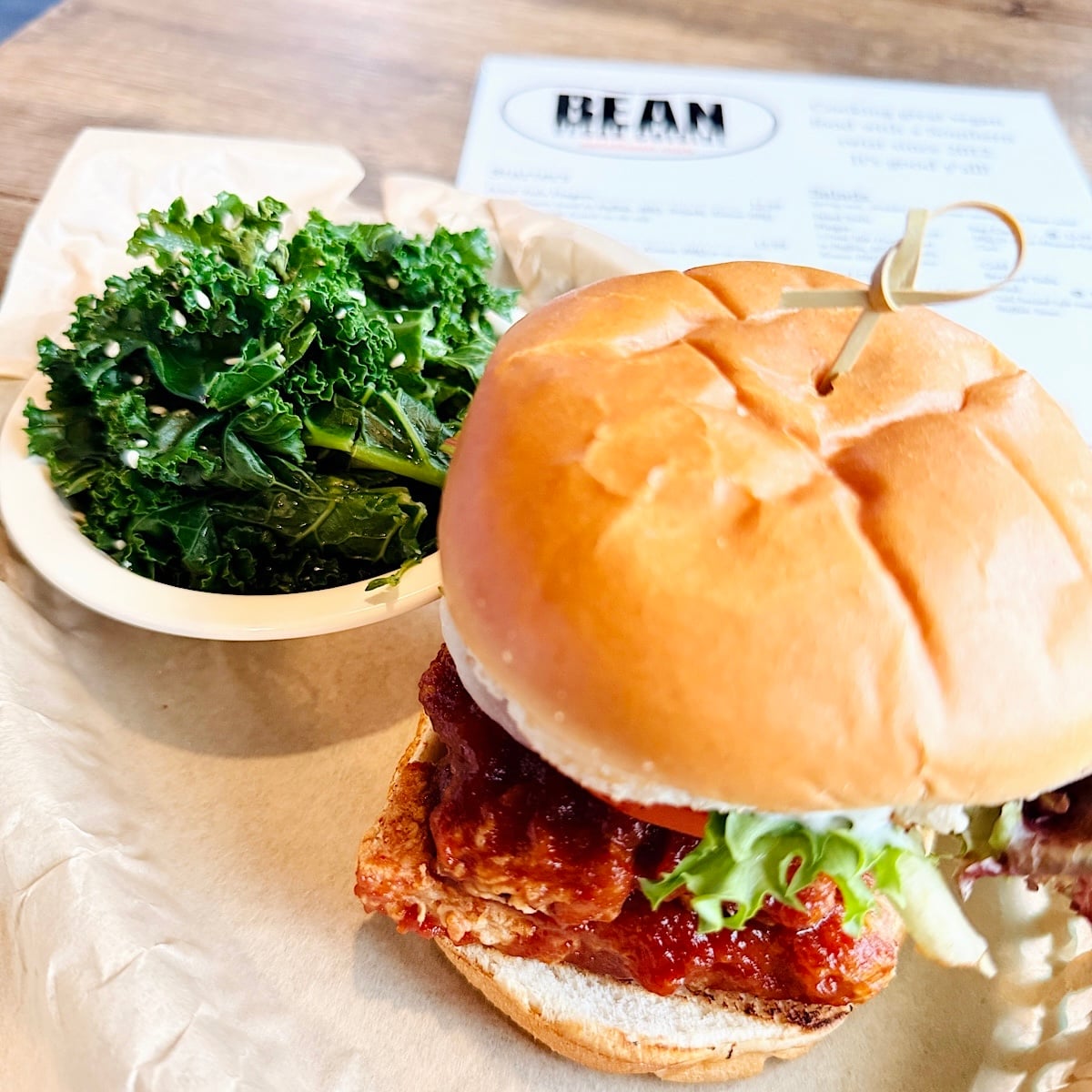 A breaded, sauced patty with lettuce on a bun sits on a tray next to a small bowl of steamed kale. A menu is blurred in the background.