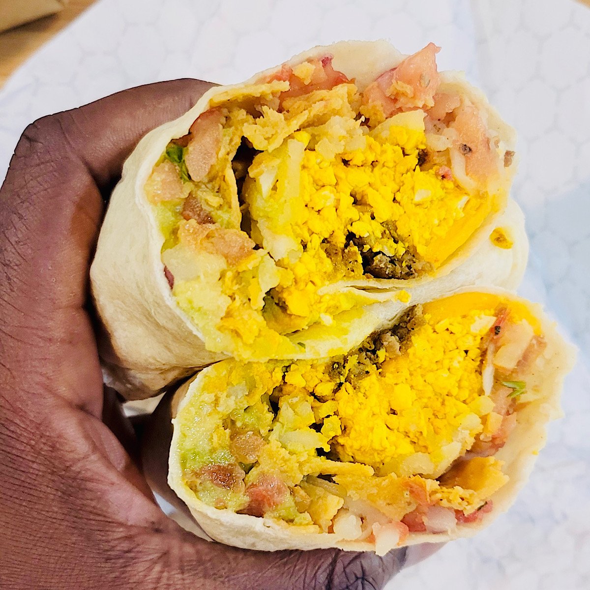A hand holds two halves of a vegan breakfast burrito filled with scrambled tofu, potatoes, and diced vegetables, wrapped in a flour tortilla.