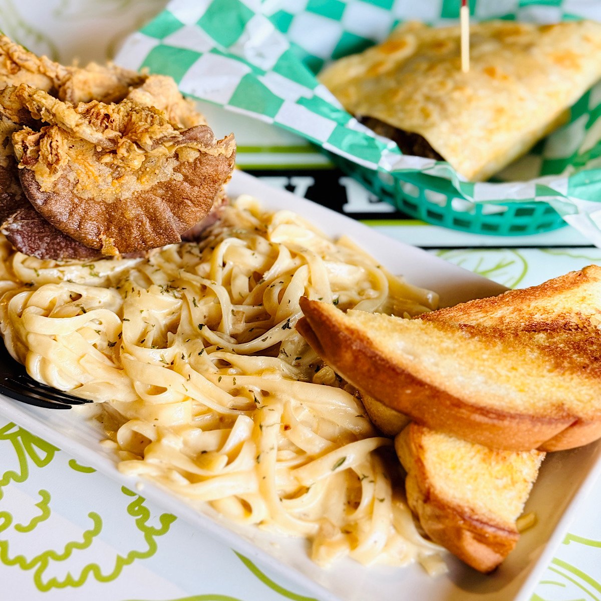 A plate of creamy vegan fettuccine Alfredo pasta topped with fried mushrooms and served with two slices of toasted garlic bread. In the background, there is a sandwich wrapped in checkered green paper.