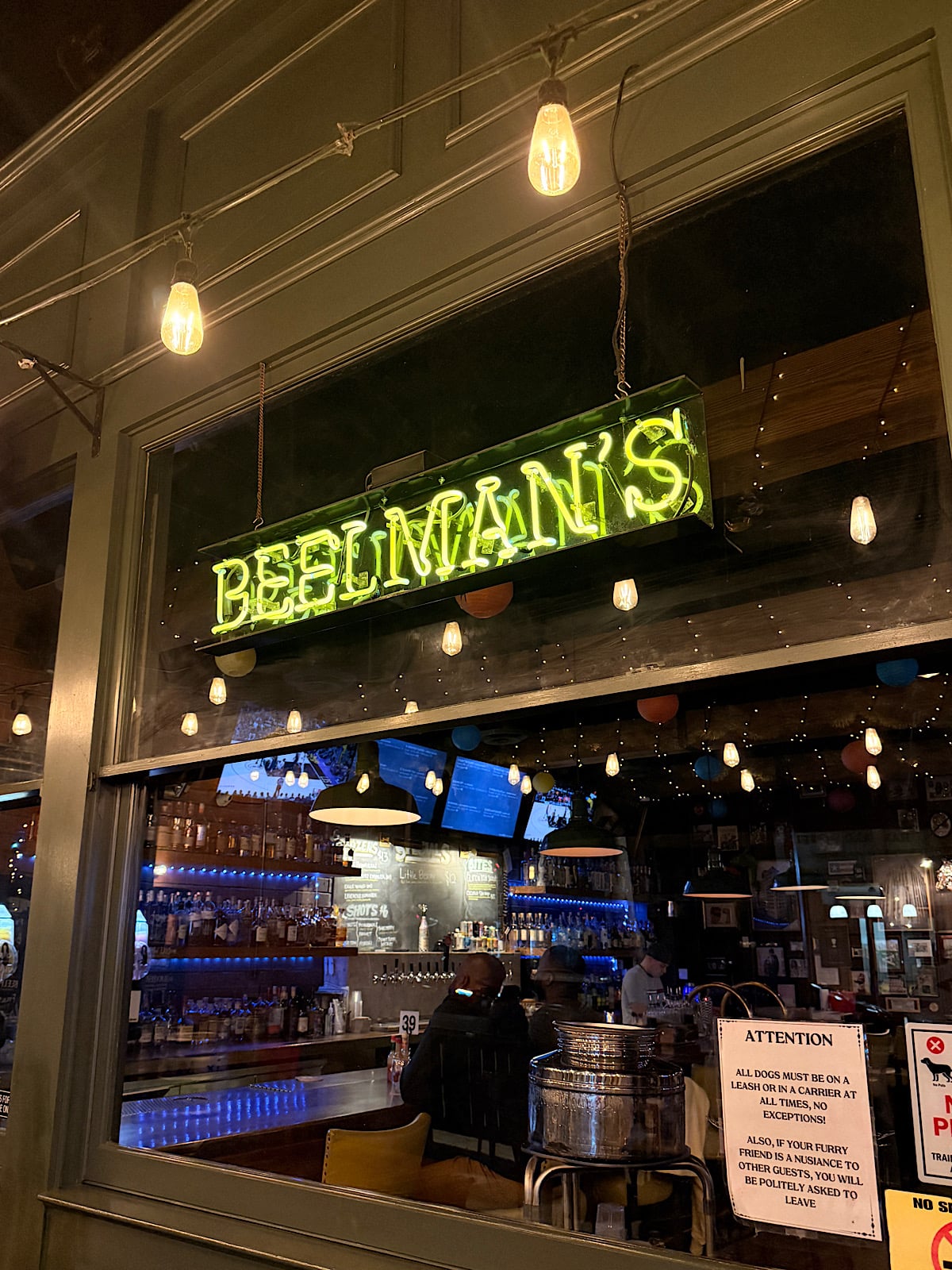 View through a window into a bar named Beelman's, with a neon sign, bottles lined up on shelves, people seated at the bar, and warm hanging lights creating a cozy atmosphere.