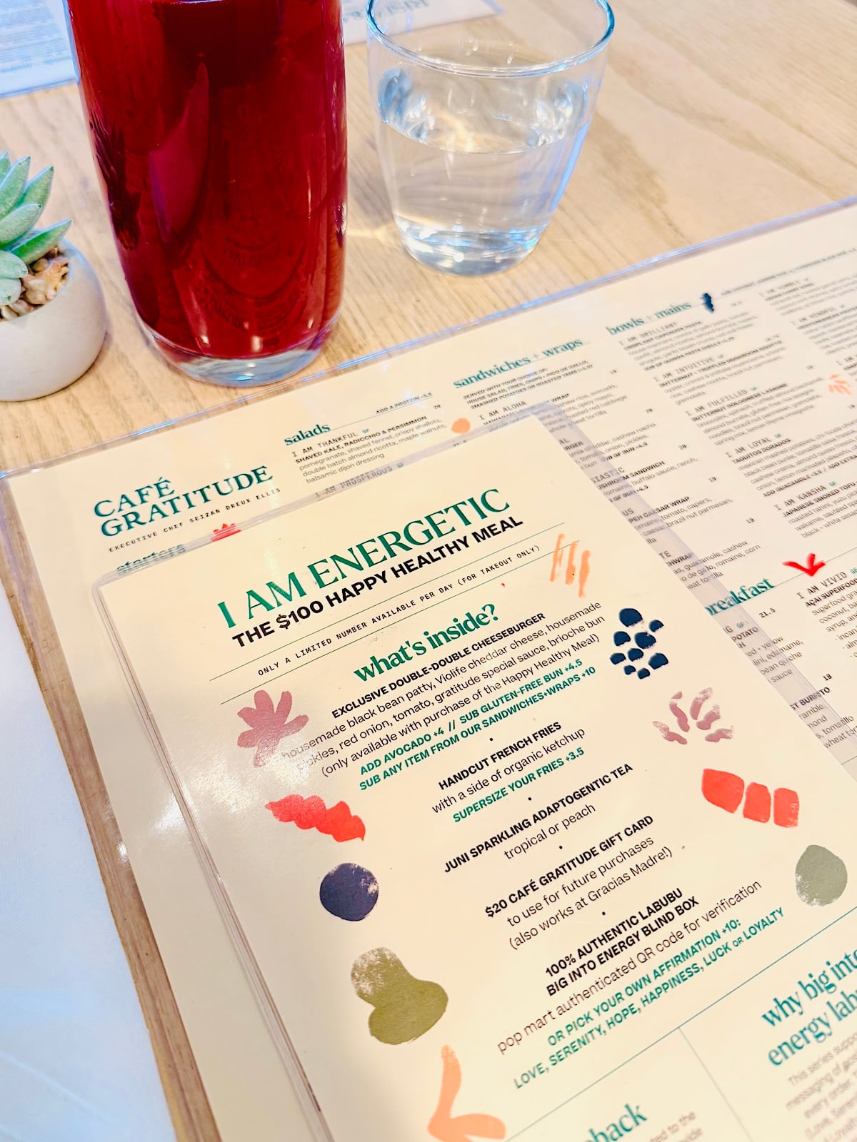 A Café Gratitude menu on a table with a featured meal titled I AM ENERGETIC, next to a glass of beet juice, a glass of water, and a small potted succulent plant.