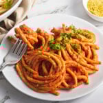 A white plate filled with thick spaghetti noodles coated in a vegan tomato sauce, garnished with chopped parsley. A fork is twirling some pasta. A bowl of grated vegan cheese or seasoning is in the background.