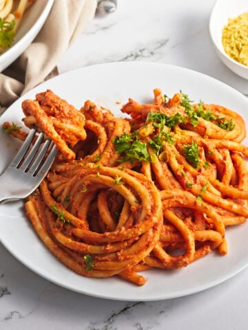 A white plate filled with thick spaghetti noodles coated in a vegan tomato sauce, garnished with chopped parsley. A fork is twirling some pasta. A bowl of grated vegan cheese or seasoning is in the background.