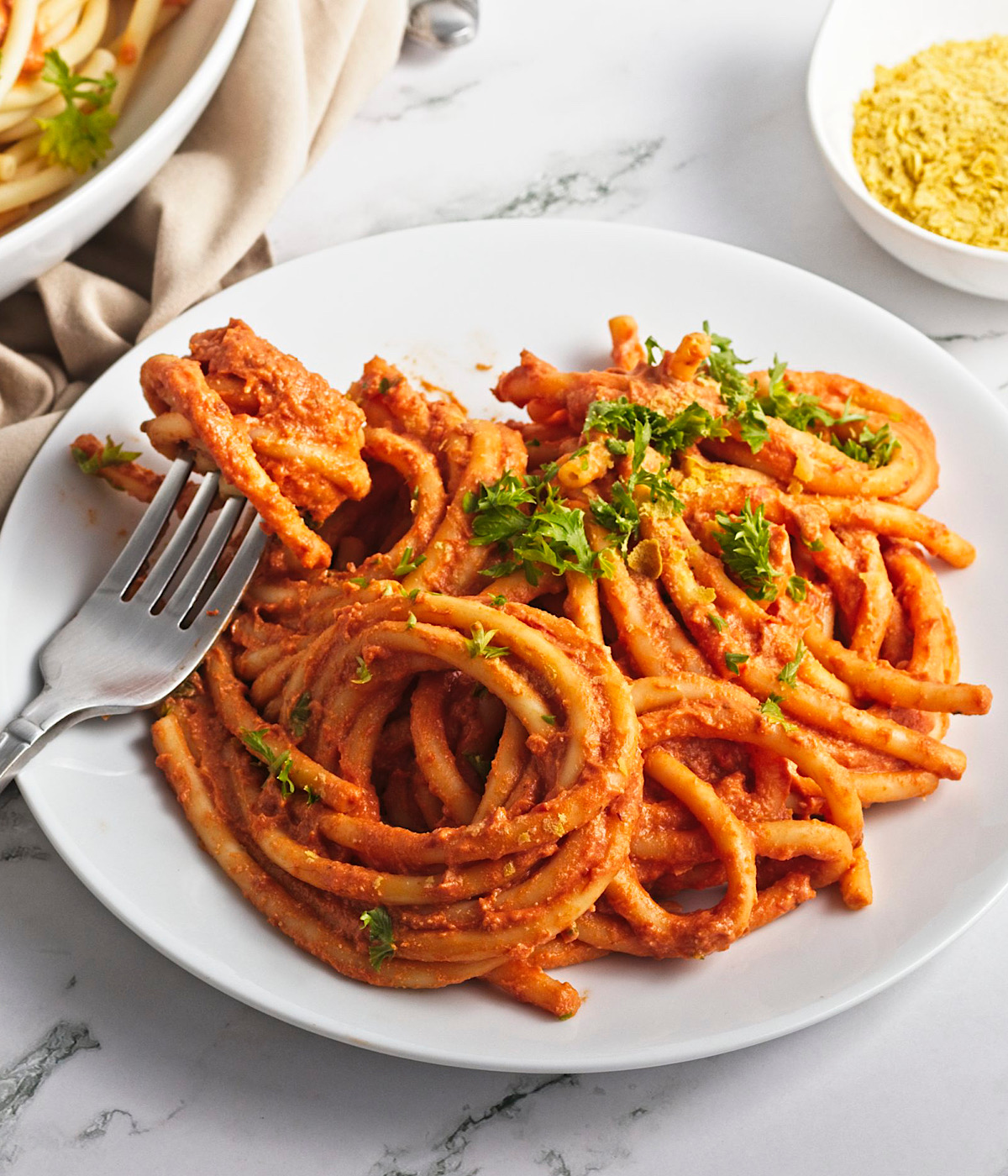 A white plate filled with thick spaghetti noodles coated in a vegan tomato sauce, garnished with chopped parsley. A fork is twirling some pasta. A bowl of grated vegan cheese or seasoning is in the background.