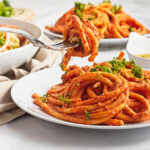 A close-up of a white plate filled with pasta noodles coated in vegan red tomato sauce, garnished with parsley. A fork lifts a twirl of pasta above the plate. Other plates of pasta are visible in the blurred background.