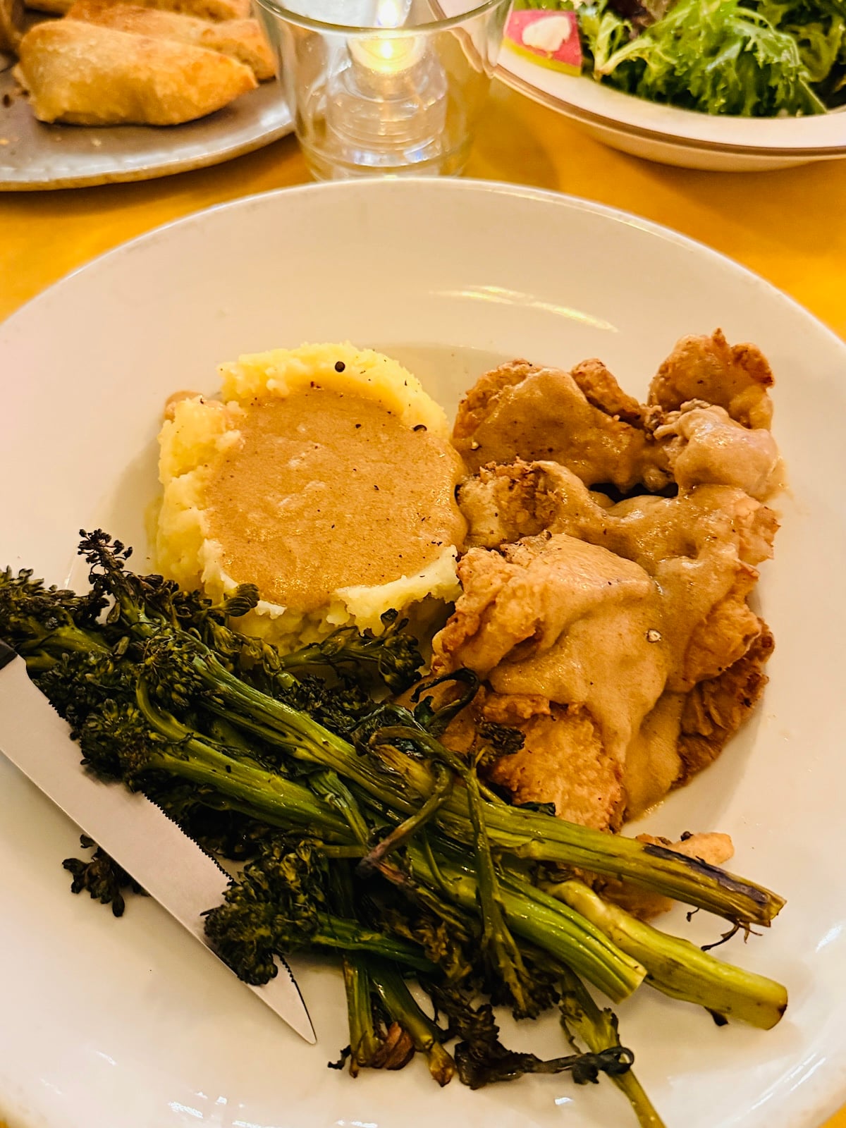 A plate of mashed potatoes with gravy, vegan fried chicken mushroom covered in gravy, and sautéed broccolini, with a knife resting on the plate; salad and bread are visible in the background.