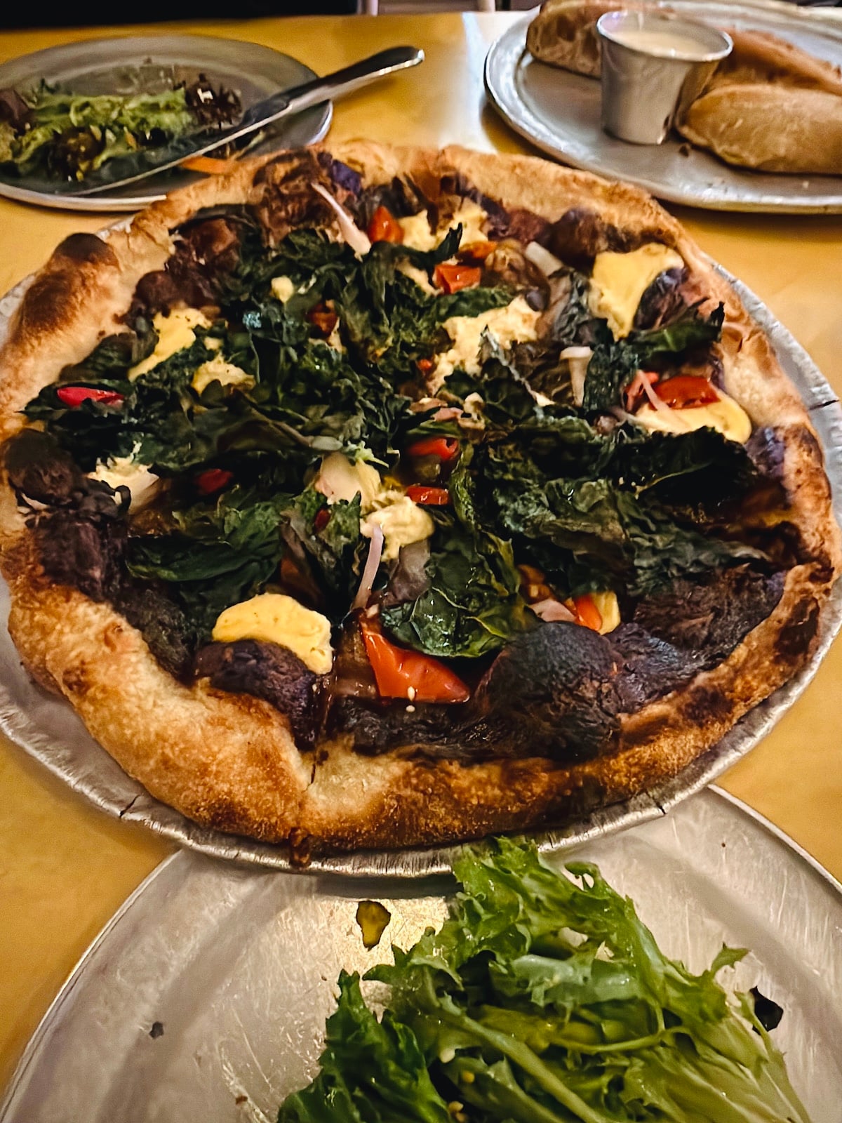 A close-up of a baked vegetable pizza topped with leafy greens, red bell peppers, onions, and dollops of cheese, served on a metal tray with a side salad and another dish in the background.