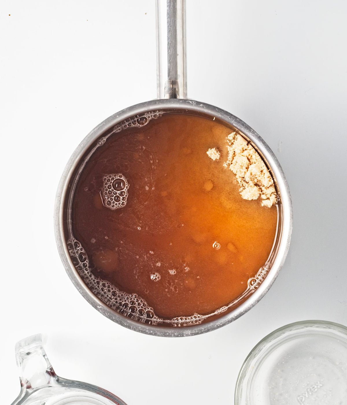 A metal saucepan filled with brown gingerbread syrup and brown sugar sits on a white surface. Nearby are a clear glass measuring cup and a clear glass bowl.