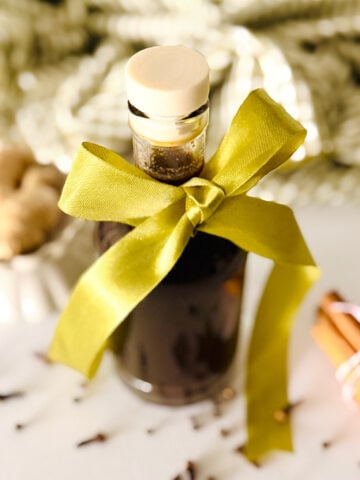A glass bottle filled with a gingerbread syrup, sealed with a cork and decorated with a large yellow-green ribbon bow, sits on a white surface surrounded by scattered spices and a blurred background.