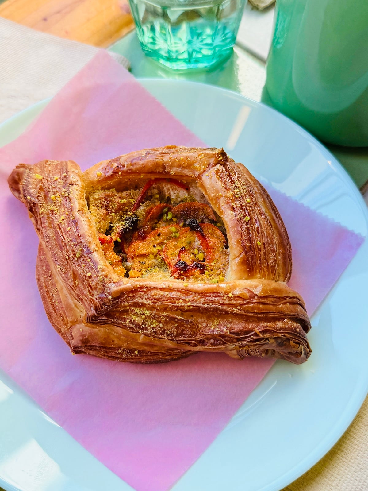 A flaky, golden-brown vegan pastry with a savory filling sits on a pink napkin atop a white plate; a green cup and a glass of water are in the background on a sunlit table.