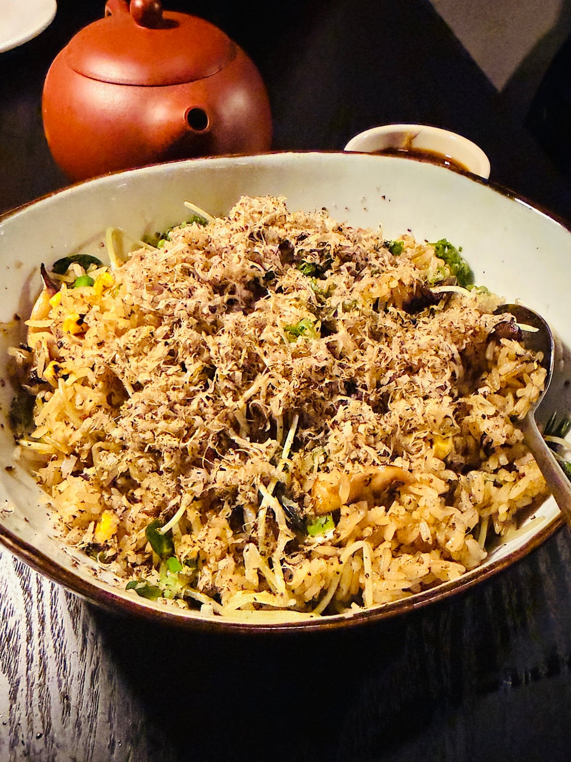 A bowl of fried rice topped with finely grated truffle, mixed with vegetables and mushrooms, with a serving spoon resting inside. A red teapot and a small sauce dish are in the background on a dark table.
