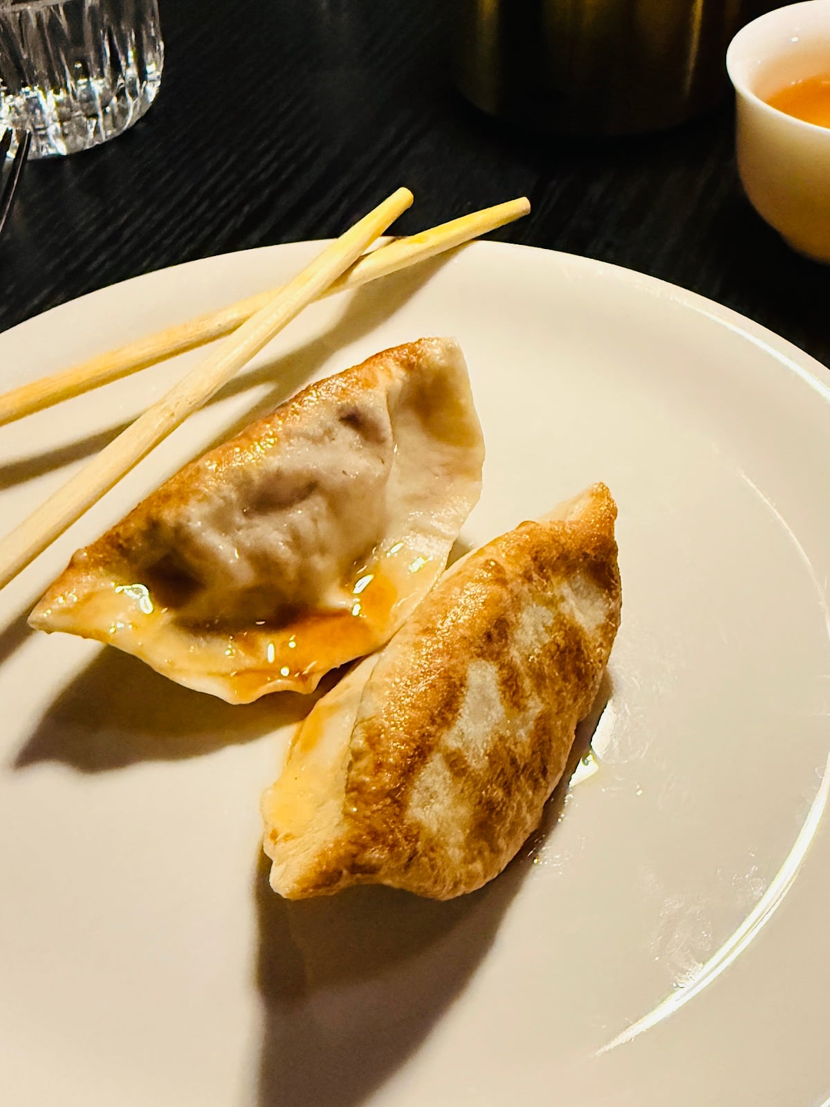 Two pan-fried dumplings with a light glaze of sauce are on a white plate, accompanied by a pair of chopsticks. A brown dipping sauce and glass of water are partially visible in the background.