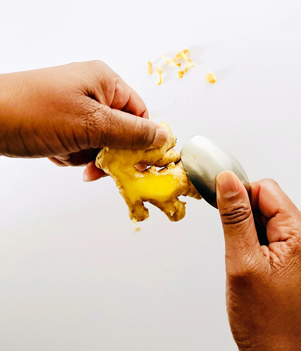 Two hands use a spoon to peel the skin off a piece of fresh ginger root, with small ginger peelings visible on a white surface.