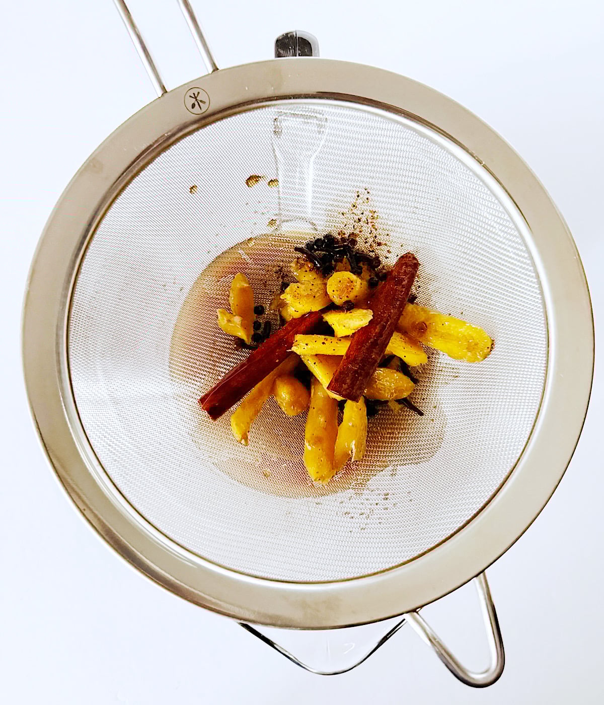 A metal strainer holds ginger slices, cinnamon sticks, and black peppercorns, viewed from above on a white background.
