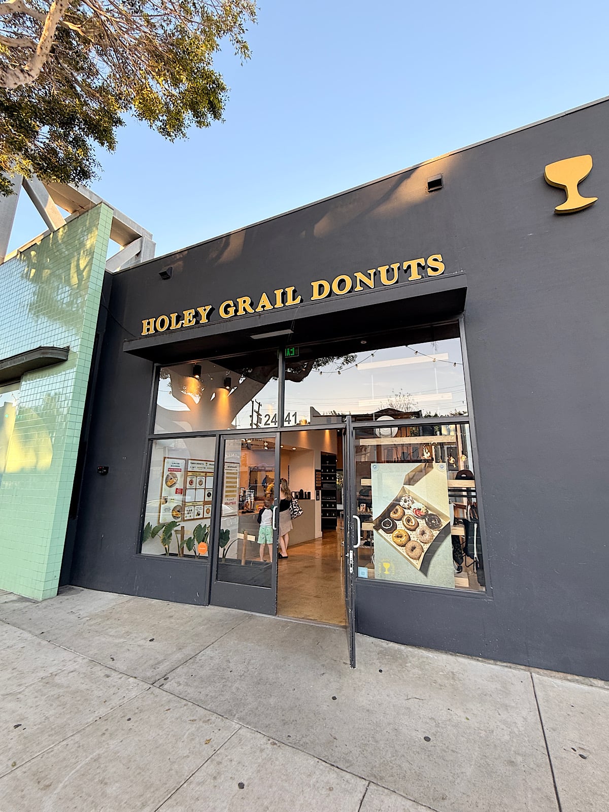 Storefront of Holey Grail Donuts with a dark gray exterior, gold lettering, and a glass door. Inside, a person is visible near the counter, and donut images are displayed on the window.