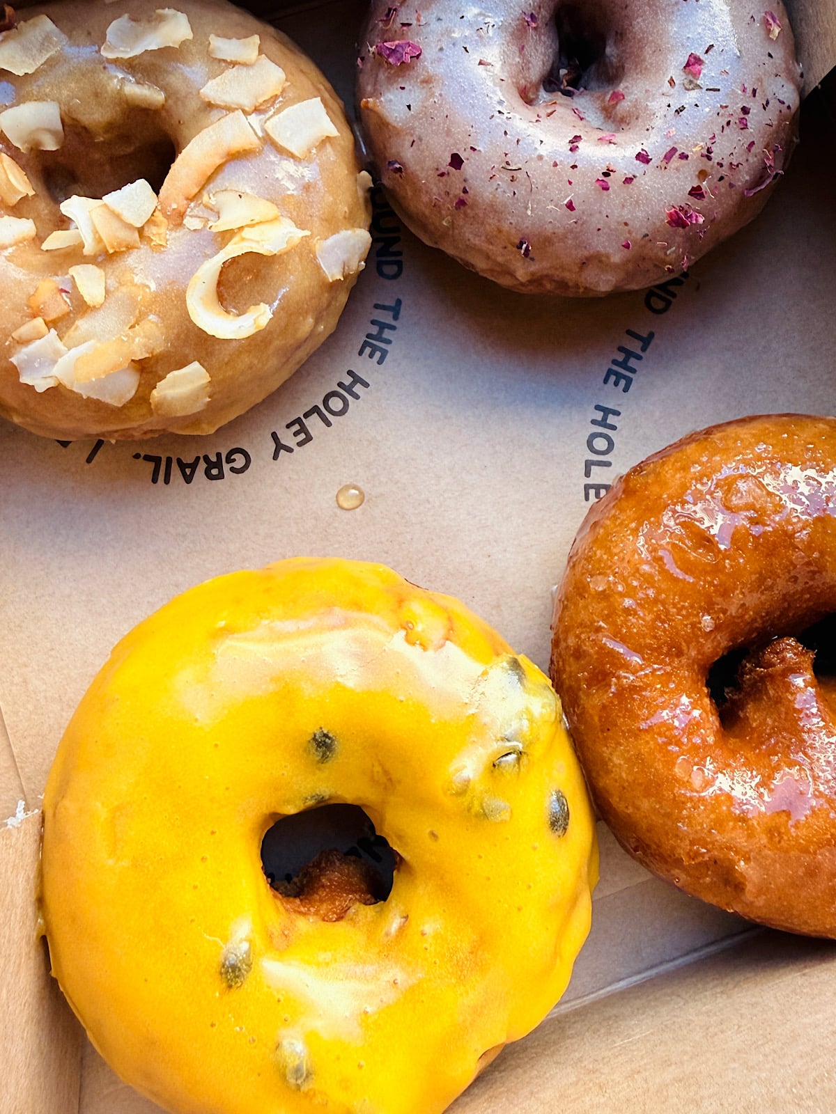 Four assorted vegan donuts in a box: one with coconut flakes, one with light brown icing and crushed red dried hibiscus flowers, one with bright yellow passion fruit icing, and one with a glossy brown glaze.