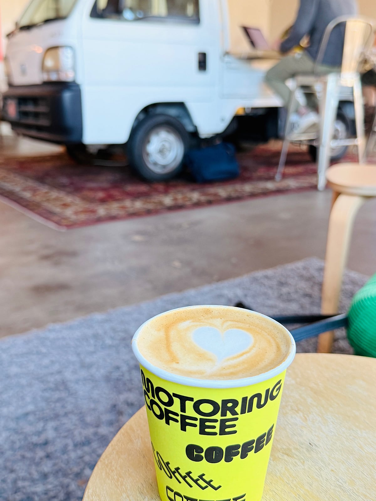 A cup of coffee with latte art in a bright yellow Motoring Coffee cup sits on a table. In the background, a small white truck is parked indoors on a rug, with a person working at a desk nearby.