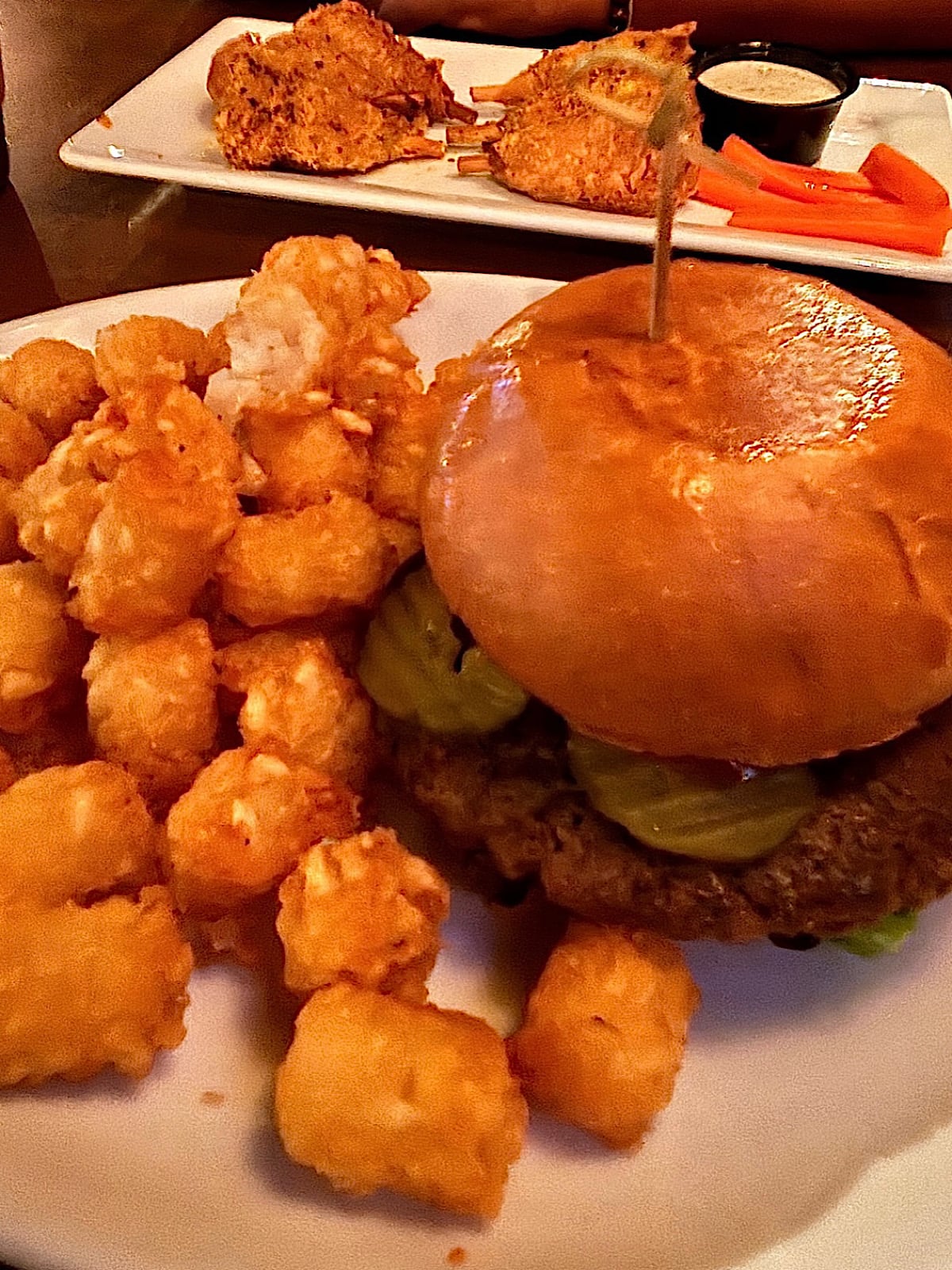 A close-up of a plate with a impossible hamburger topped with pickles and a side of crispy tater tots. In the background, there are fried food items, carrot sticks, and a cup of dipping sauce on another plate.