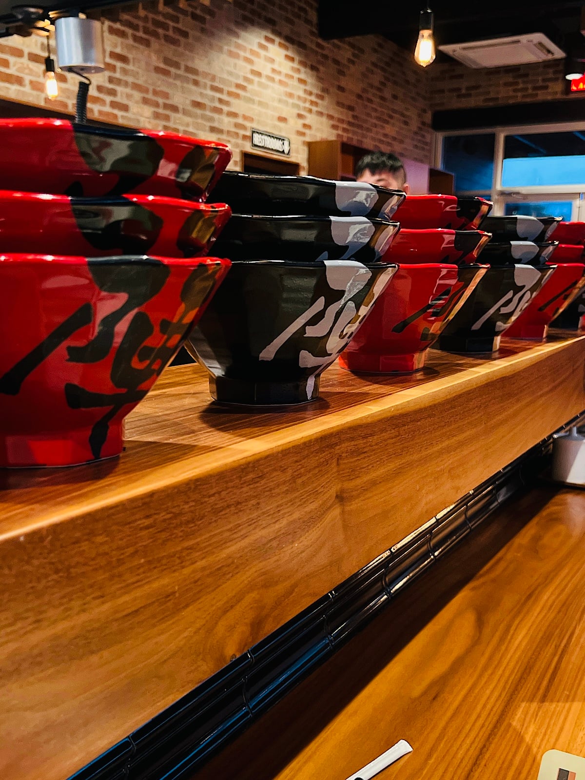 A row of stacked red and black ramen bowls with calligraphic designs sits on a wooden counter inside a restaurant with brick walls and warm lighting.
