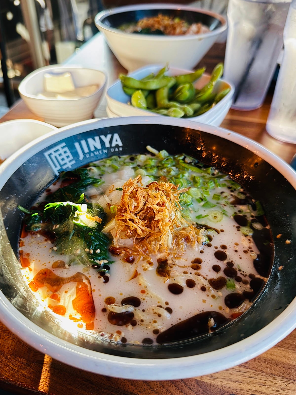 A bowl of ramen topped with fried onions, scallions, and black garlic oil from JINYA Ramen Bar, with sides of edamame, tofu, and drinks visible in the background on a wooden table.