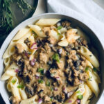 A bowl of penne pasta topped with a creamy vegan mushroom sauce, garnished with chopped red onions, green herbs, and fresh rosemary beside the dish on a white napkin.