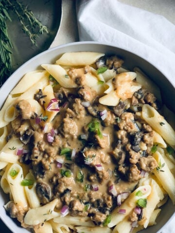 A bowl of penne pasta topped with a creamy vegan mushroom sauce, garnished with chopped red onions, green herbs, and fresh rosemary beside the dish on a white napkin.