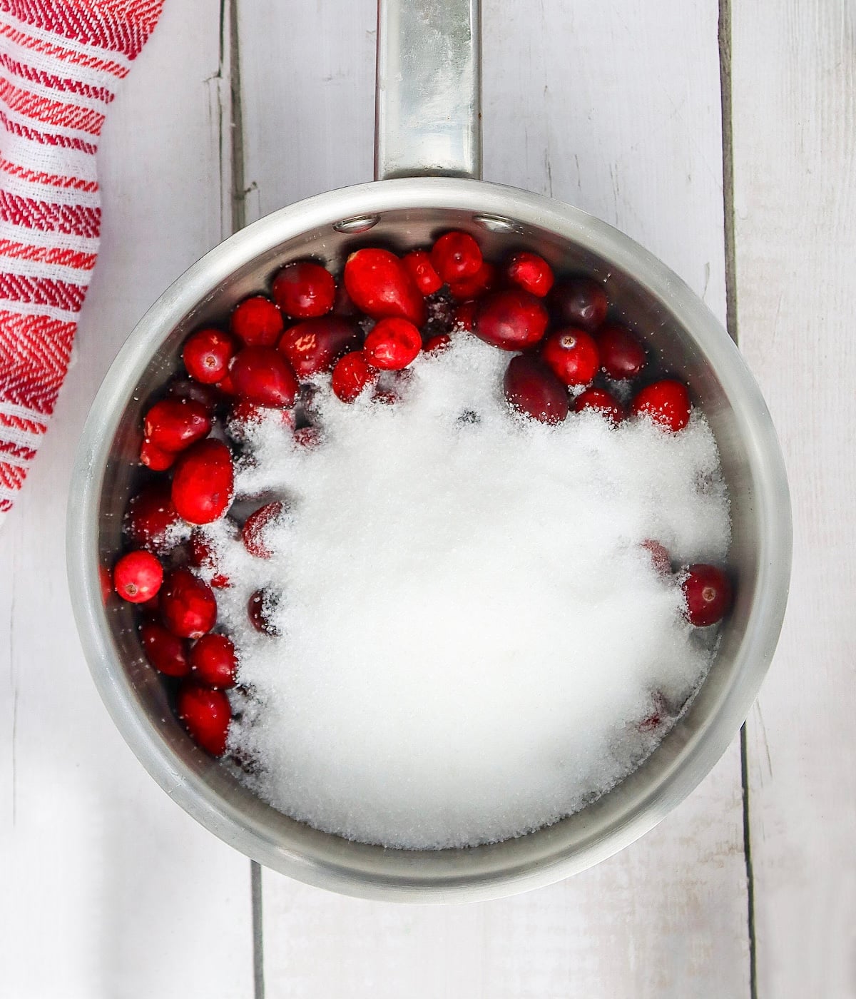 A saucepan filled with fresh cranberries and white sugar on a white wooden surface next to a red and white striped towel.