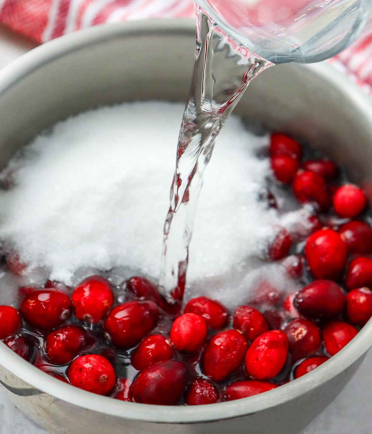 A pot filled with fresh cranberries and sugar is being topped with a stream of water poured from a glass measuring cup. A red and white towel is visible in the background.