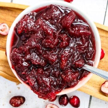 A white bowl filled with chunky cranberry sauce sits on a wooden tray. Whole and halved fresh cranberries are scattered around the bowl, and a spoon is placed inside the sauce.