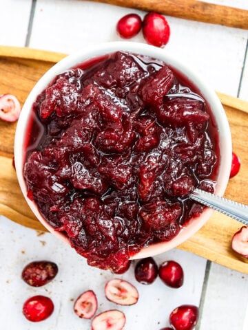 A white bowl filled with chunky cranberry sauce sits on a wooden tray. Whole and halved fresh cranberries are scattered around the bowl, and a spoon is placed inside the sauce.
