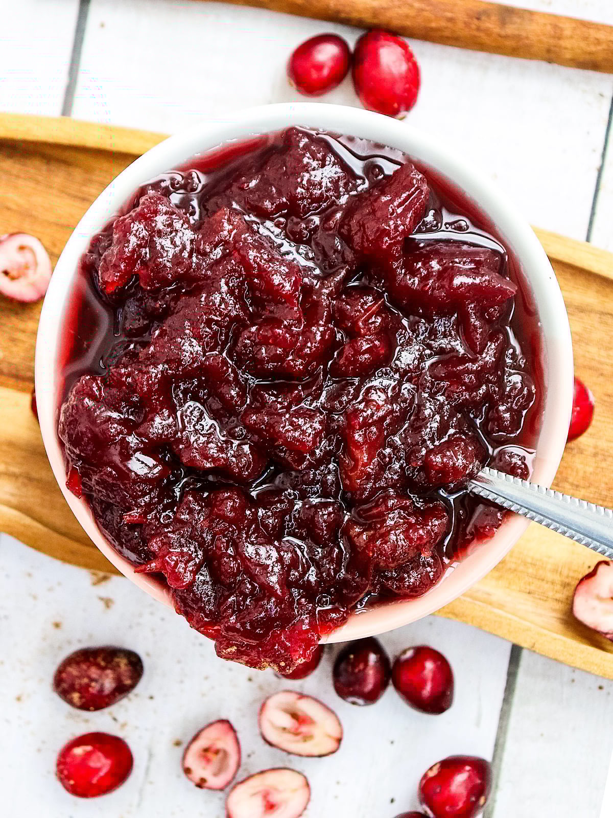 A white bowl filled with chunky cranberry sauce sits on a wooden tray. Whole and halved fresh cranberries are scattered around the bowl, and a spoon is placed inside the sauce.