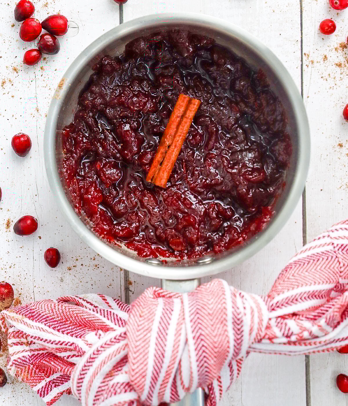 A saucepan filled with cranberry sauce and a cinnamon stick, surrounded by scattered cranberries on a white wooden surface, with a red and white striped towel wrapped around the handle.