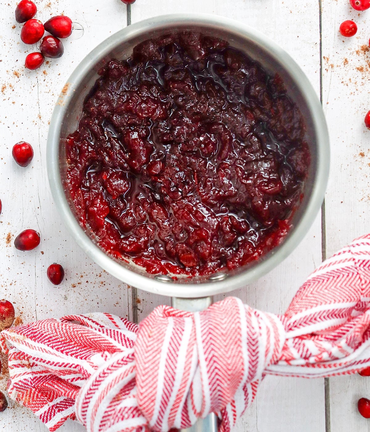 A saucepan filled with homemade cranberry sauce sits on a white wooden surface, surrounded by fresh cranberries. The handle is wrapped with a red and white striped kitchen towel.