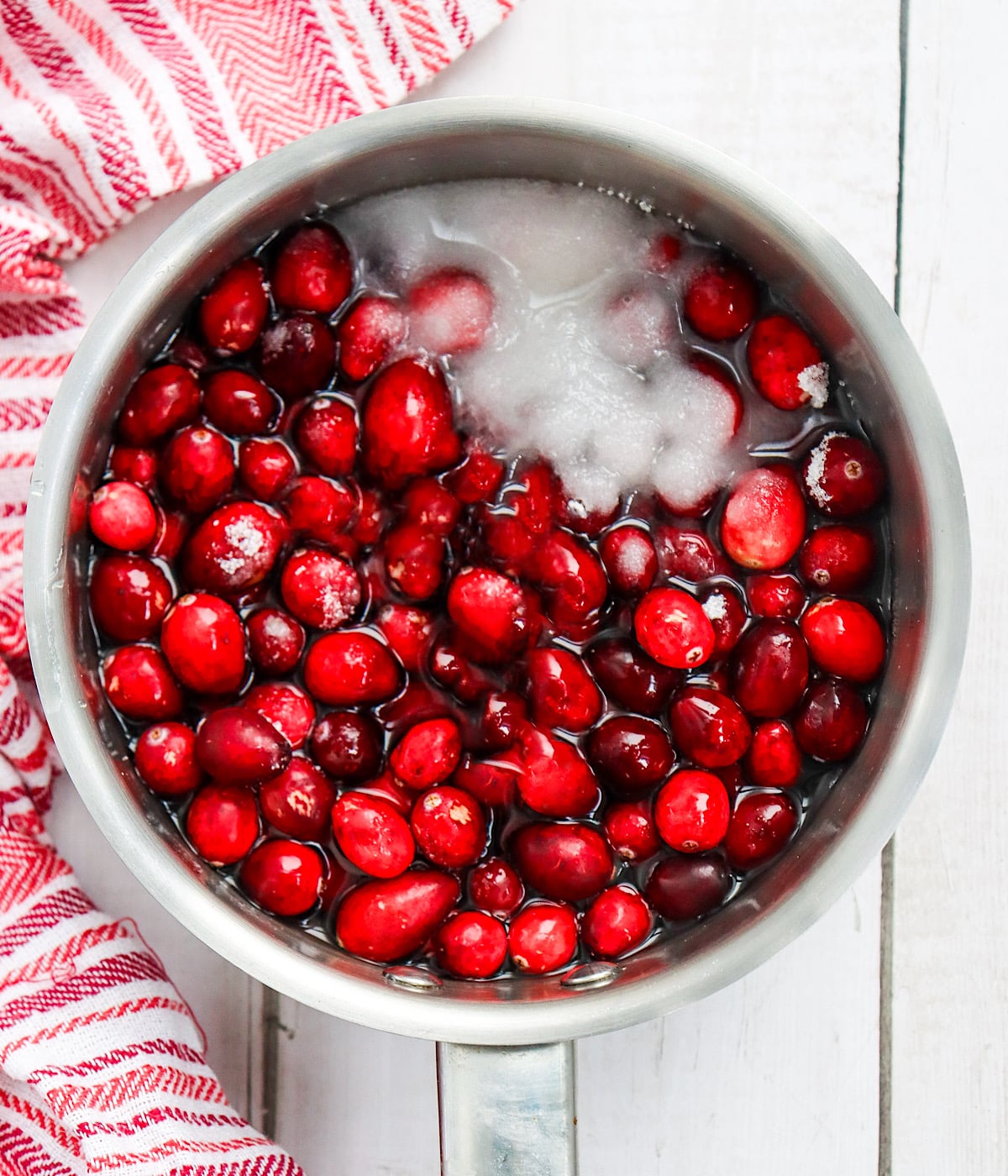 A saucepan filled with fresh cranberries, water, and sugar sits on a white wooden surface next to a red and white striped towel.