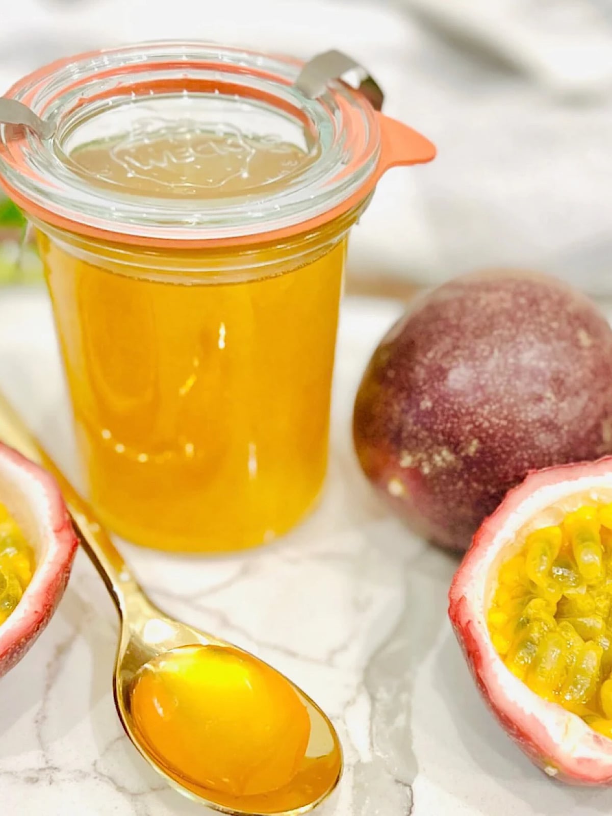 A glass jar filled with bright yellow passion fruit syrup sits on a marble surface, next to a gold spoon with syrup, a whole passion fruit, and a halved passion fruit showing its seeds.