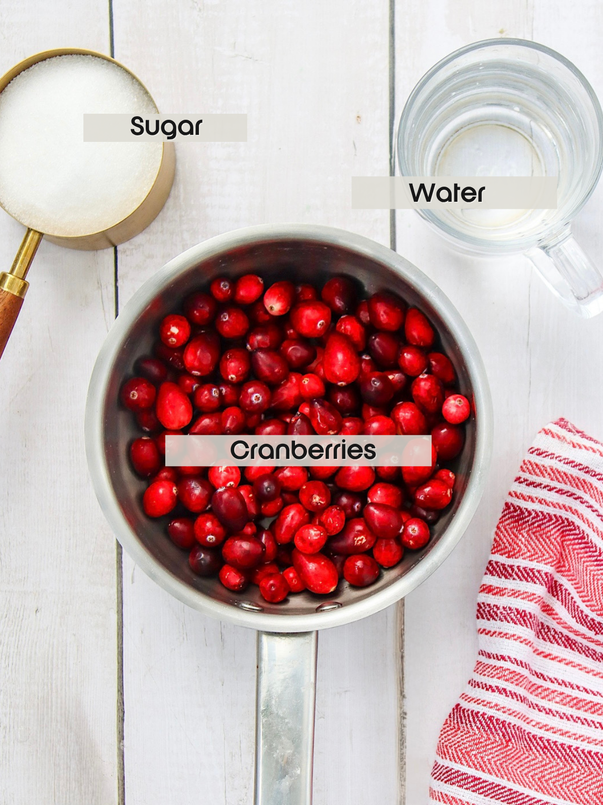 Overhead view of a saucepan filled with fresh cranberries, next to a measuring cup of sugar and a glass of water on a white wooden surface, with a red-striped towel nearby. Labels identify sugar, water, and cranberries.